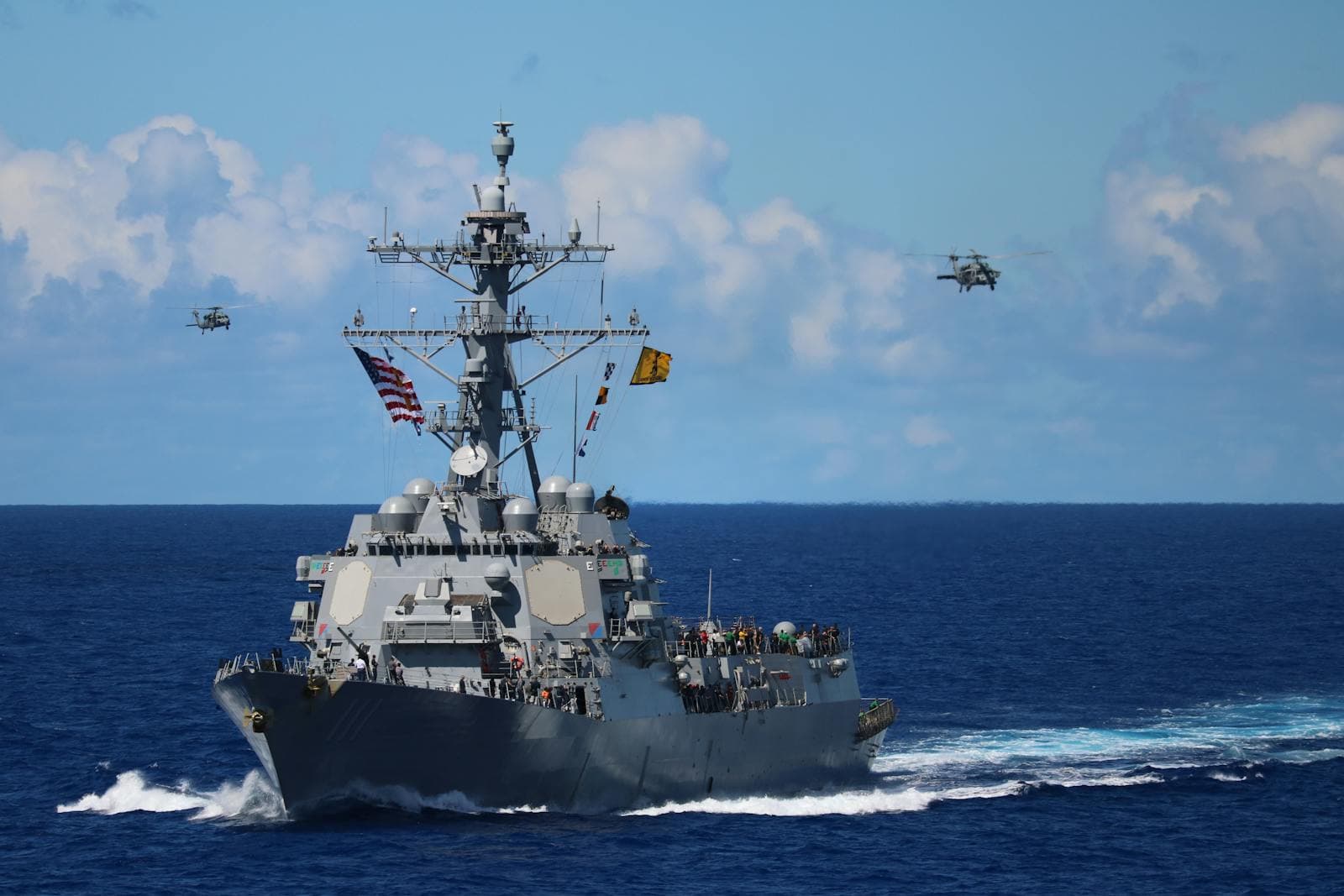 US Navy destroyer at sea with helicopters in formation overhead.